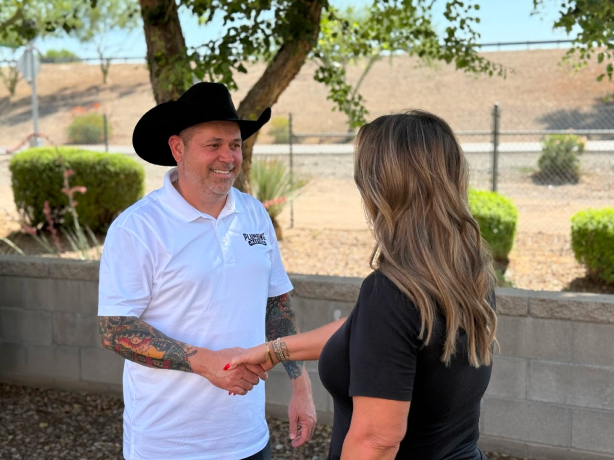 A man in a cowboy hat and white polo shirt shakes hands with a woman outdoors near a low wall and greenery.