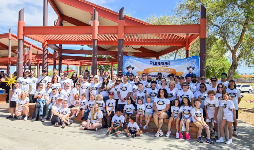 A large group of people in matching shirts pose for a photo outdoors in front of a banner for a plumbing company anniversary event.