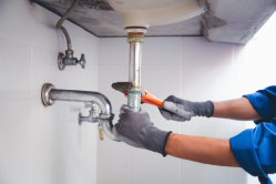 A person wearing gloves uses a wrench to adjust plumbing pipes under a sink.